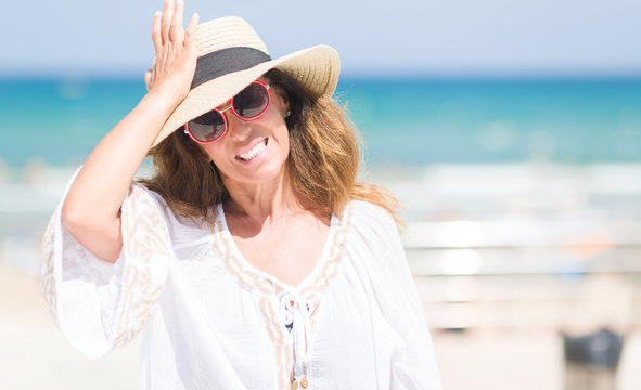 Middle age brunette woman wearing summer hat on vacations by the beach stressed with hand on head, shocked with shame and surprise face, angry and frustrated. Fear and upset for mistake.
