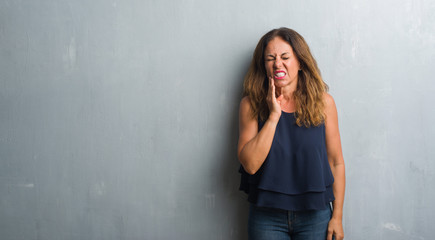 Middle age hispanic woman standing over grey grunge wall touching mouth with hand with painful expression because of toothache or dental illness on teeth. Dentist concept.