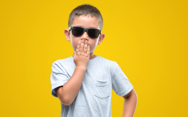 Dark haired little child wearing sunglasses cover mouth with hand shocked with shame for mistake,...