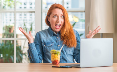 Redhead woman using computer laptop eating fruit at home very happy and excited, winner expression celebrating victory screaming with big smile and raised hands