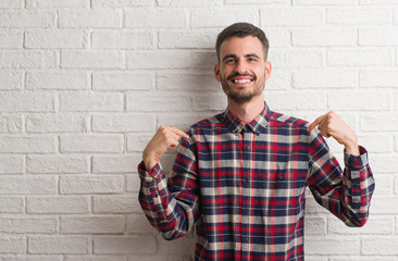 Young adult man standing over white brick wall looking confident with smile on face, pointing...