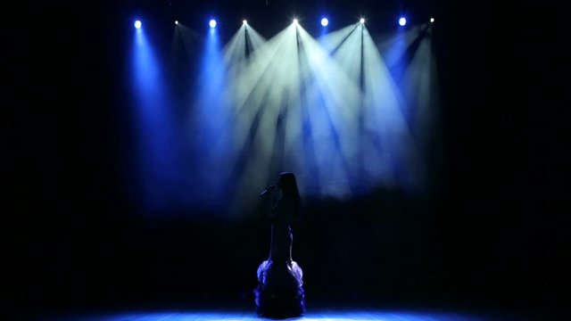 Silhouette of a luxurious singer in an evening gown on a stage in the dark. The girl sings a song on an empty stage in the light of a blue spotlight.