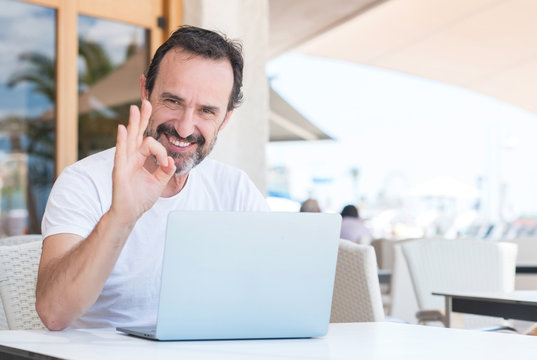 Handsome senior man using laptop at restaurant doing ok sign with fingers, excellent symbol