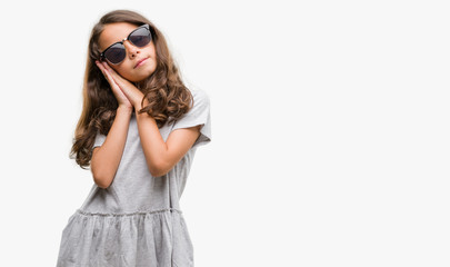 Brunette hispanic girl wearing sunglasses sleeping tired dreaming and posing with hands together while smiling with closed eyes.