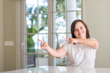 Down syndrome woman at home smiling and looking at the camera pointing with two hands and fingers to the side.