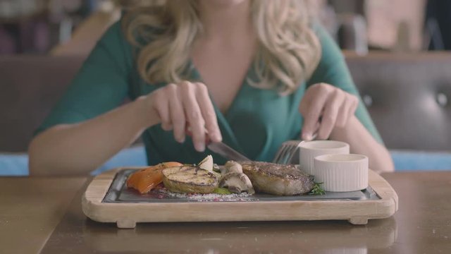 The food blurred concept: attractive blonde lady eating delicious steak in restaurant slow motion. Only hands, woman holding fork and knife and tasting meat with grill vegetables on the wooden plate