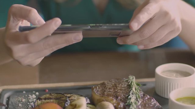 The food photo concept: female hands holding smartphone and making a picture of restaurant dish on the wooden plate. Delicious meat steak with grilled vegetables, sauces, grains of salt and herbs
