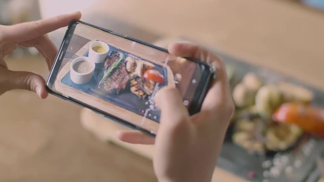 The smartphone screen and food photo concept. Woman's hands making a few picture of delicious steak with vegetables at wooden tray in the grill restaurant from the top, pushing on the touchscreen