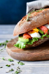 French sandwich from a cereal baguette with avocado, salmon, egg and lettuce leaves on a wooden plate. wooden table and dark background, selective focus and copy space