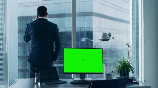 Confident Businessman in His Office Making Phone Call, Personal Computer on His Desk Shows Green Chroma Key Screen. In the Window Panoramic Big City View.