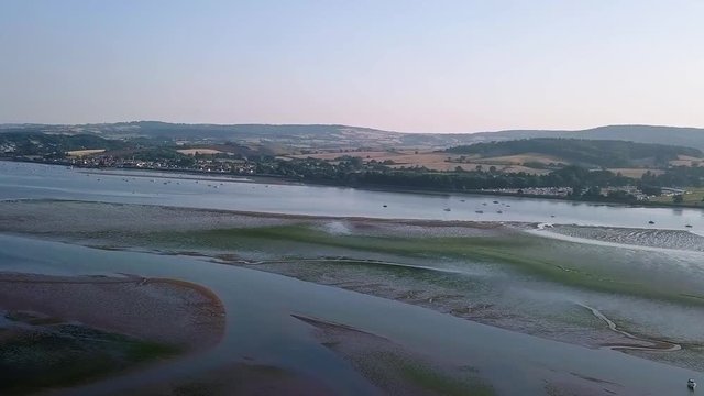 Flying over the docked ships and sail boat off of the shore of Lympstone England. The boats and land are at a distance but well visible. This location is visited by tourists daily.