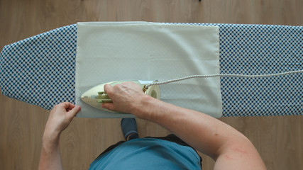 TOP VIEW: Young adult man ironing a white pillowcase on an ironing board at home