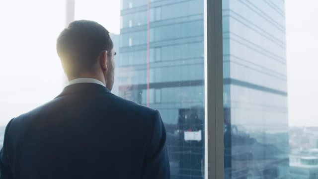 Medium Shot of Successful Businessman wearing a Suit Standing in His Office, Contemplating Next Big Business Deal, Looking out of the Window. Big City Business District Panoramic Window View.