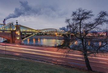 Moscow, Russia. View of the Bogdan Khmelnitsky (Kievsky) Pedestrian Bridge and the Rostov embankment. In the foreground lawn and a large tree. In the background, the smoke from the pipes and cloud sky