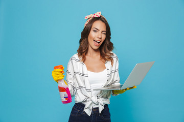 Portrait of european housewife 20s in yellow rubber gloves holding detergent sprayer and laptop during cleaning, isolated over blue background