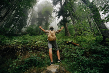 male tourist in shorts, a hat and with a backpack in the misty forest of a hand in the side gesture of admiration.