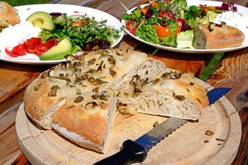 Homemade Italian rosemary, green olive and onion Focaccia bread on a wooden board with salad to the rear.