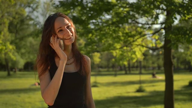 Smiling young woman stand at the park outdoors talk by phone She looks happy nad cheerful. 4k slow motion.
