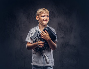 Happy blonde schoolboy dressed in a white t-shirt, holds full digital set for entertainment at a studio. Isolated on dark textured background.