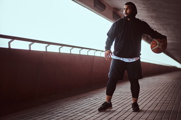 Portrait of a dark-skinned guy dressed in a black hoodie and sports shorts playing basketball on a footway under bridge.