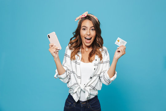 Image of emotional caucasian girl 20s wearing headband laughing while holding smartphone and credit card, isolated over blue background