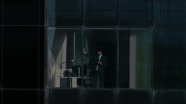 Aerial View Footage: From Outside into Office Building with Businessman Using Mobile Phone and Standing Next to the Office Window. Shot on RED EPIC-W 8K Helium Cinema Camera.