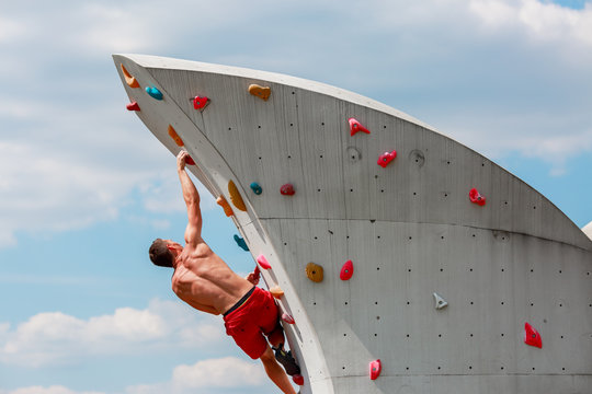Photo from back of young sporty man in red shorts hanging on wall for rock climbing against blue sky with clouds