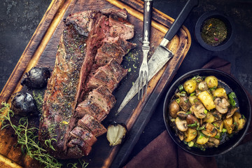 Traditional barbecue aged marinated saddle of venison with fried potatoes and mushroom as top view on a cutting board