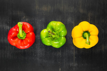 Top view of three fresh bright bell peppers on black rustic background. Shot from above of green, yellow and red paprika vegetables on dark wood table