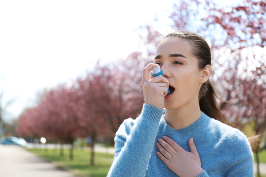 Young woman using inhaler near blooming trees. Allergy concept