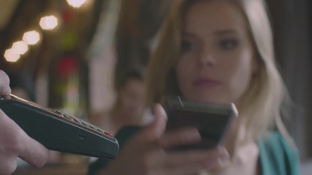 Blurred concept: caucasian woman finish a drinking cup of coffee and using the smartphone for contactless payment in restaurant. Waiter' hand holding the terminal and lady pay the bill for drinks