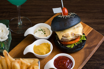 Black burger on board on dark wooden table background. Black burger with meat patty, cheese, tomatoes, mayonnaise, french fries in a paper cup. Modern fast food lunch