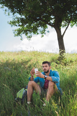 Naklejka na ściany i meble male tourist using smartphone and eating sandwich on summer meadow with tree