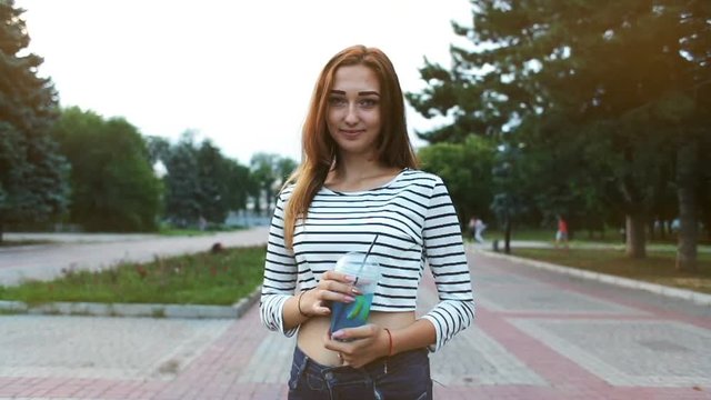 stylish beautiful girl in tops and jeans drinks a summer drink walking through the city park