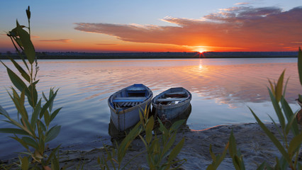 Two boats stand at the shore of a calm river against the background of a bright rising sun