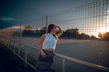 Fototapeta na wymiar Attractive young blonde woman in stylish white blouse and skirt posing near the wire fence on evening. Dark stormy clouds. Urban background