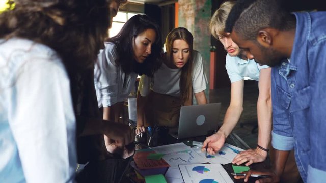 Group of young people colleagues is discussing charts and graphs standing around table in modern office. Business, teamwork and professionals concept.