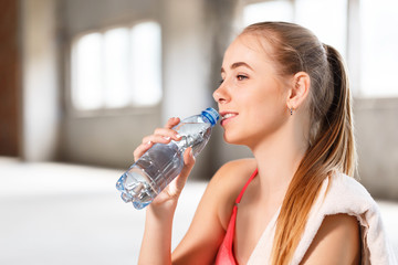 Athlete happy fitness girl with perfect body shape drinking from plastic bottle of water after workout on the sport mat in the sunny loft gym