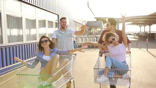 Boys pushing shopping carts with girls at sunset near shopping mall. Young people having fun. They very happy and cheerful