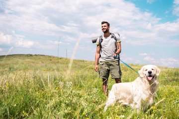 traveler with backpack walking with golden retriever on summer meadow with cloudy sky