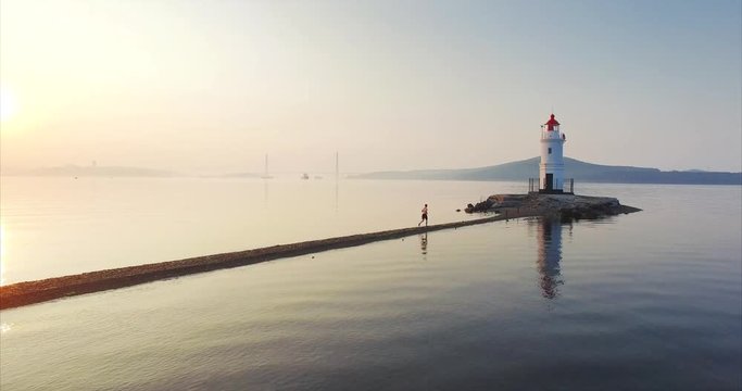 Beautiful aerial view of man in shorts running fast along the Tokarevsky spit to lighthouse. Flying above the sandbeach. Calm morning sea at amazing sunrise. Vladivostok, Russia
