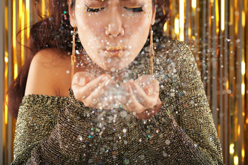 Crop view of gorgeous young ethnic brunette woman with shimmer makeup in party clothes blowing bright sparkling silver glitter at camera standing over blurred golden studio background with closed eyes