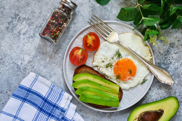 Classic breakfast: toast with avocado, egg and vegetables on a concrete background.