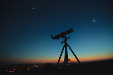 Silhouette of a telescope with stars on the sky above the city lights.