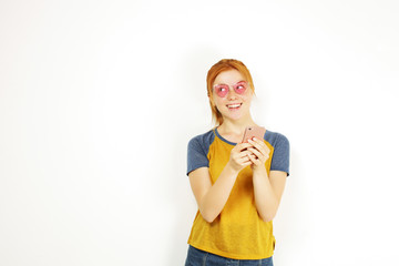 Young attractive natural redhead woman smiling at smartphone screen, using mobile phone, wearing baseball shirt & cat eye glasses. White copy space background. Female using online dating app concept