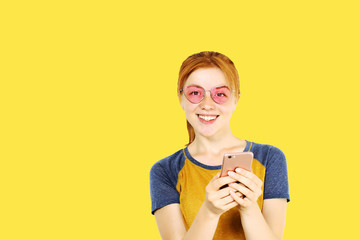 Young attractive natural redhead woman smiling at smartphone screen, using mobile phone, wearing baseball shirt & cat eye glasses. Pink copy space background. Female using online dating app concept