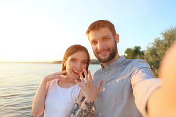 Happy couple taking selfie with engagement ring near river on sunny day