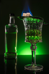 glass and bottle of absinthe with burning sugar on spoon on reflective surface and dark green background