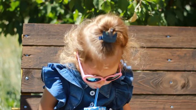 Child girl in sunglasses drinks smoothies in the park on a sunny windy day