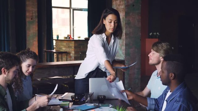 Confident businesswoman is giving papers with visual information to her creative team and talking at meeting in office. People, teamwork and documents concept.